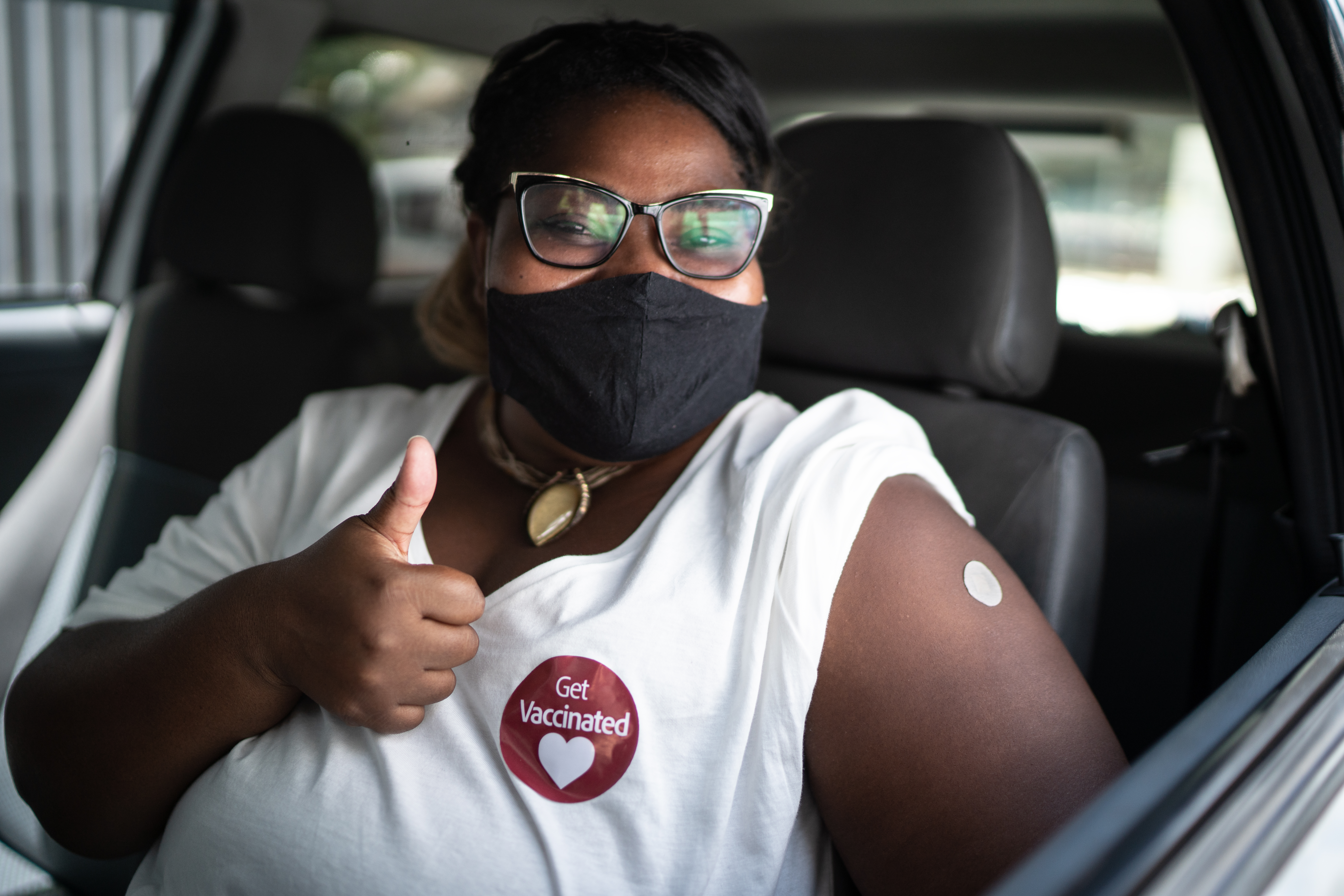 Woman sitting in driver's side of car proudly displaying vaccination sticker