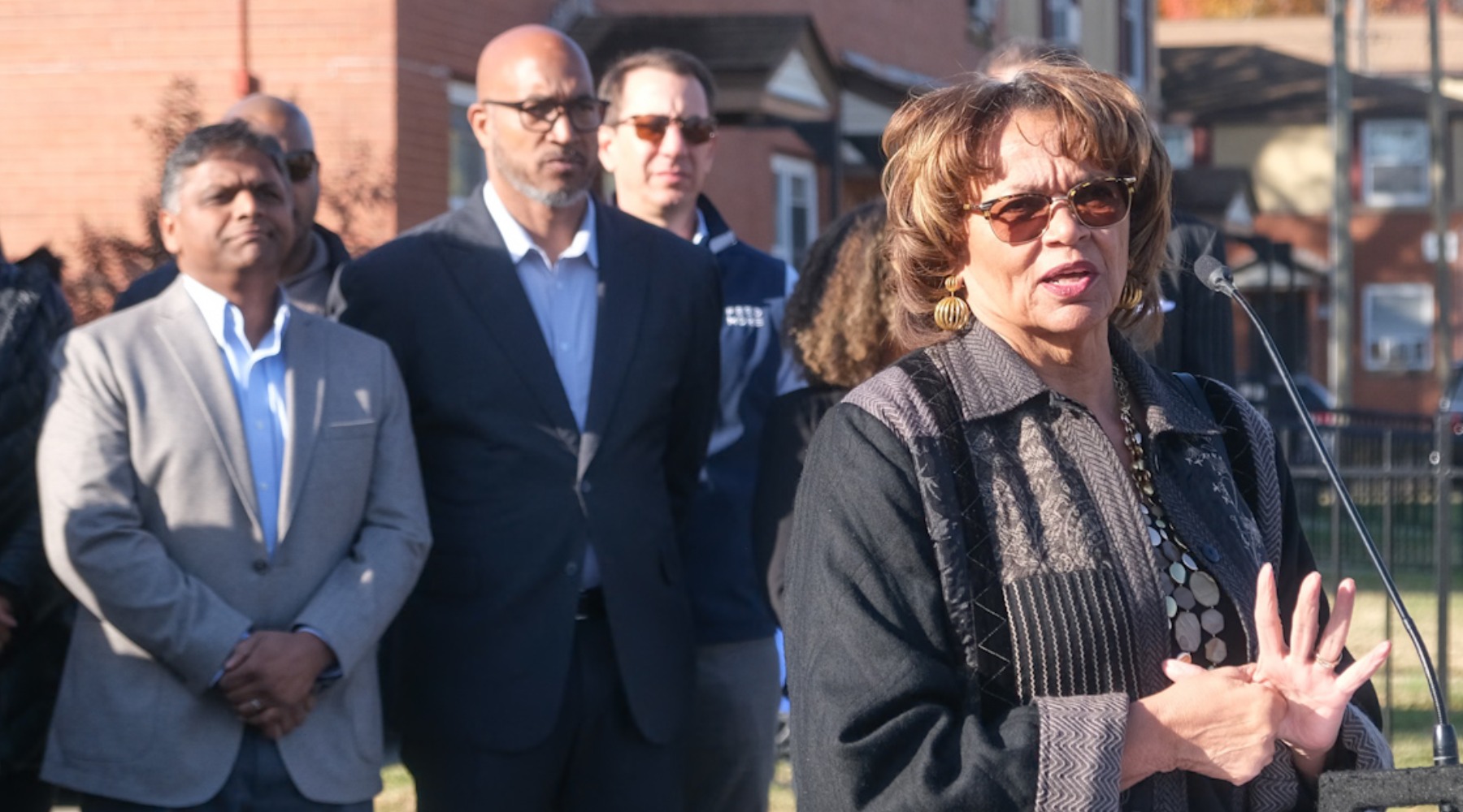 Speaker at an outdoor event, hands clasped at a microphone, with attendees in business attire behind; brick houses and a fence in the sunny background.