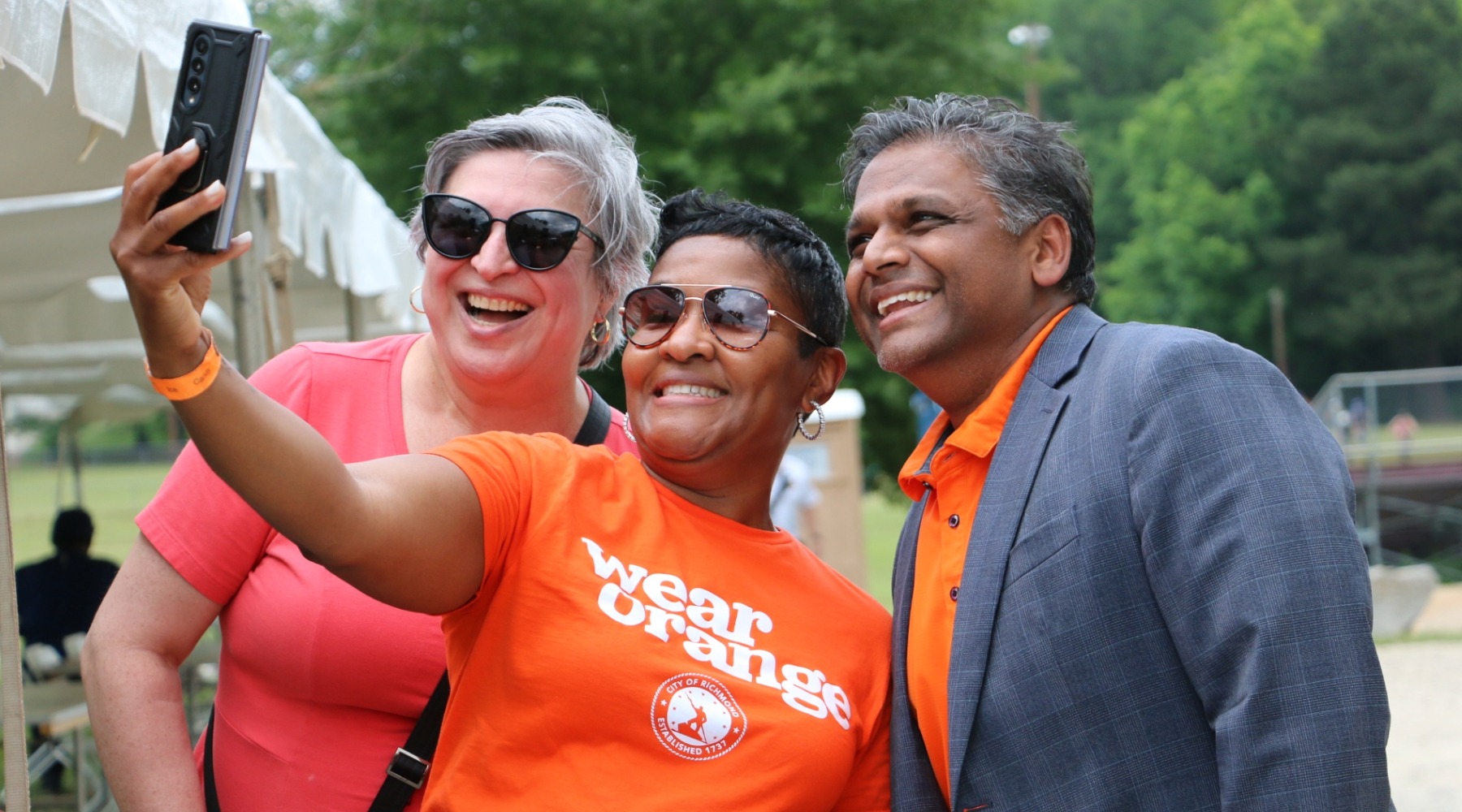 Participant at a gun violence prevention event in an orange “wear orange” shirt extends an arm to take a selfie with two others beside a white event tent; leafy park scenery behind.