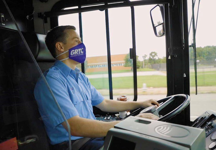 This image contains a person with short hair wearing a purple mask with GRTC logo driving a bus. 