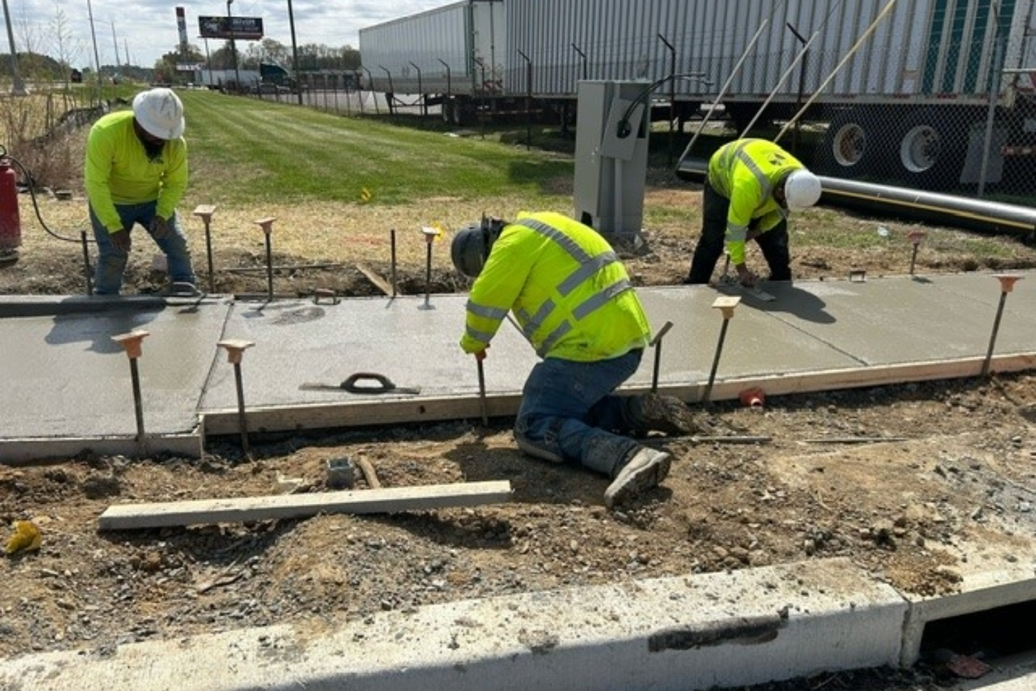 Three workers in neon green shirts pour concrete for a new sidewalk.