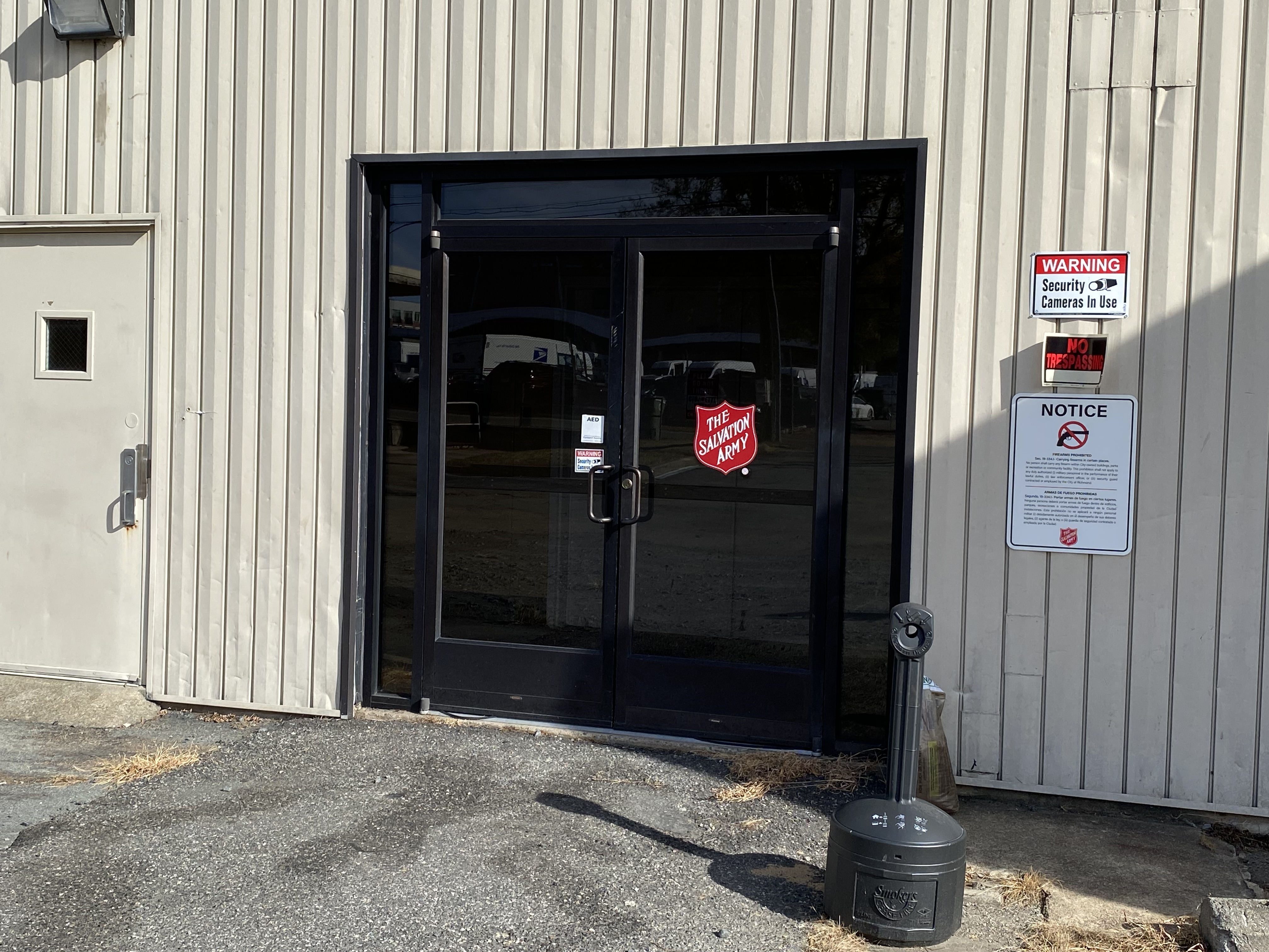 The Salvation Army building entrance with dark glass double doors, security camera warning sign, and no trespassing notice on a metal-sided wall.