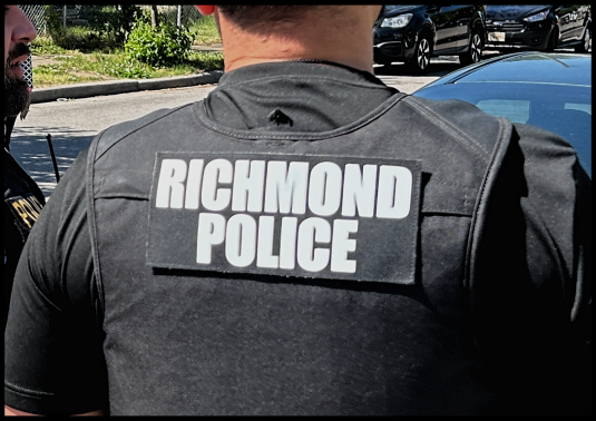 Back view of a person wearing a vest with a large “RICHMOND POLICE” patch, standing near parked cars on a sunny street.